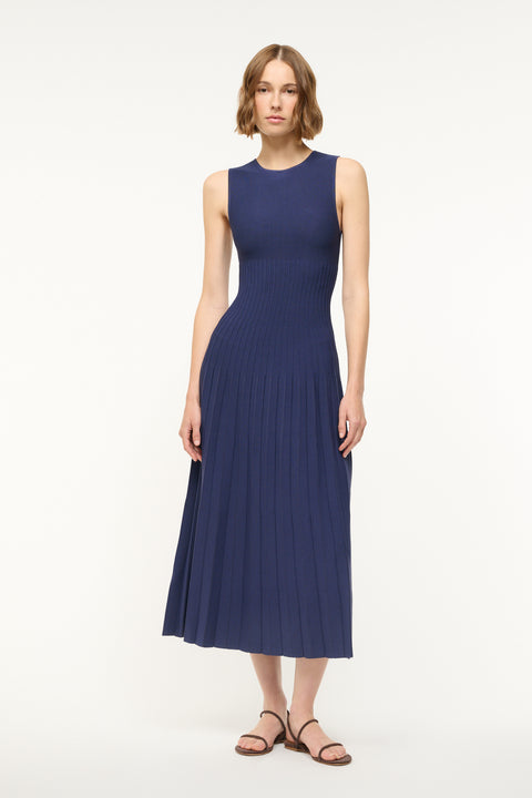 Woman wearing a navy knit midi dress that features a high neckline and flattering ribbing in the torso with a pleated skirt. 