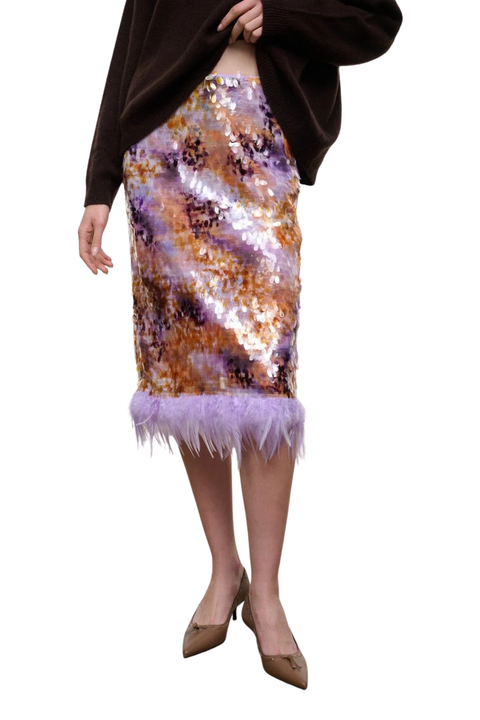 A model wearing a purple and gold sequined pencil skirt with a lavender feather trim, paired with a dark brown top and beige pointed-toe heels, shown against a plain white background.