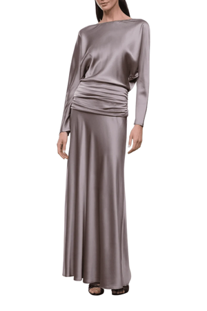 A woman modeling a floor-length satin gown in a muted taupe-gray color. The dress features a boat neckline, long sleeves, and a draped ruched waist detail, shown against a plain white background.
