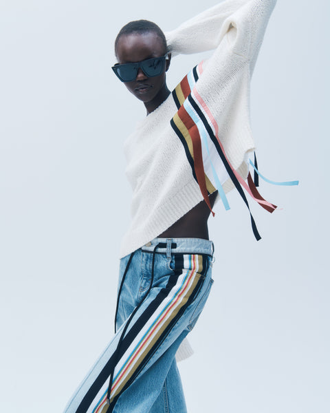 Person wearing a white sweater with colorful stripes and blue jeans on a light gray background