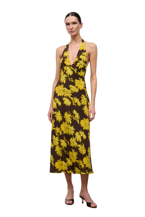 A woman modeling a sleeveless halter-neck midi dress with a bold yellow tropical floral print on a dark brown background, paired with black heeled sandals, set against a plain white studio background.