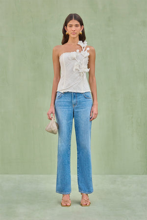 Woman wearing a linen mesh strapless top with an over-the-shoulder sculptural flower detail paired with blue jeans. 