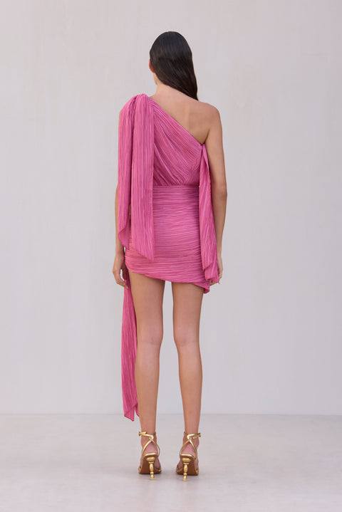 Back view of a woman wearing a pink asymmetrical mini dress made from a plisse fabric with three draping details. 