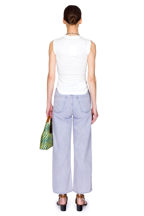 Woman wearing a white sleeveless top and high-rise, light blue jeans, holding a yellow and blue bag on a white background from the back