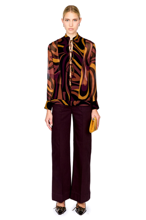 A full-length studio shot of a model wearing a long-sleeve silk shirt with an abstract wavy pattern in brown, gold, and black, paired with dark plum wide-leg trousers and holding a small yellow clutch.