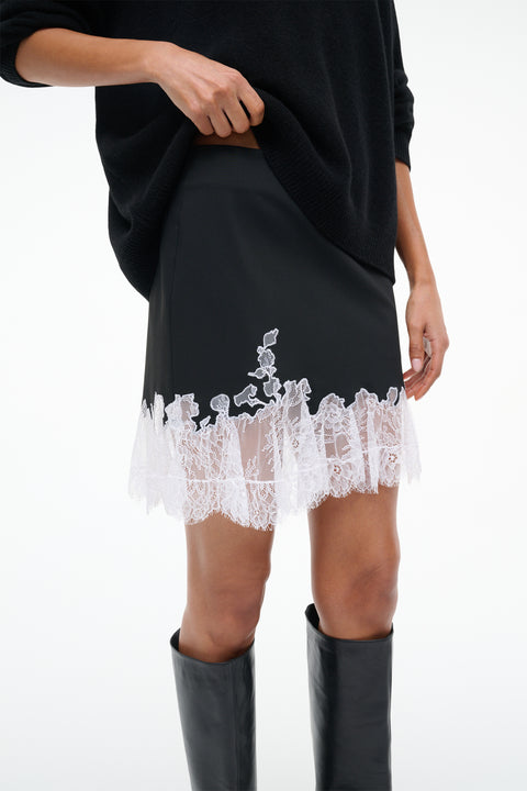 Side view of a woman wearing a black mini skirt with white lace hem, paired with a black sweater and black knee-high boots.