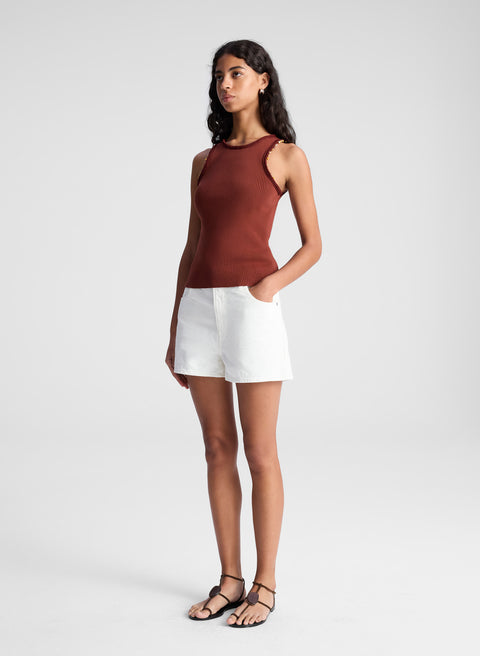 Woman facing sideways wearing a rust-colored sleeveless top and white shorts on a light gray background