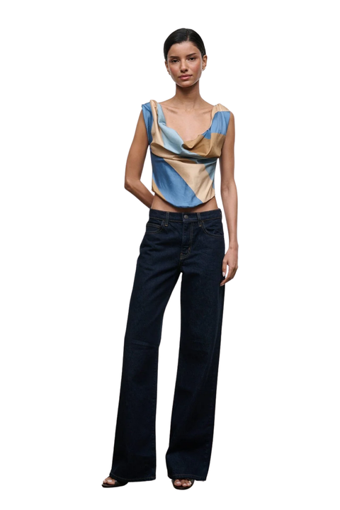 A model stands against a plain white background wearing a sleeveless draped top with blue, beige, and brown panels, paired with dark wide-leg jeans and black sandals.