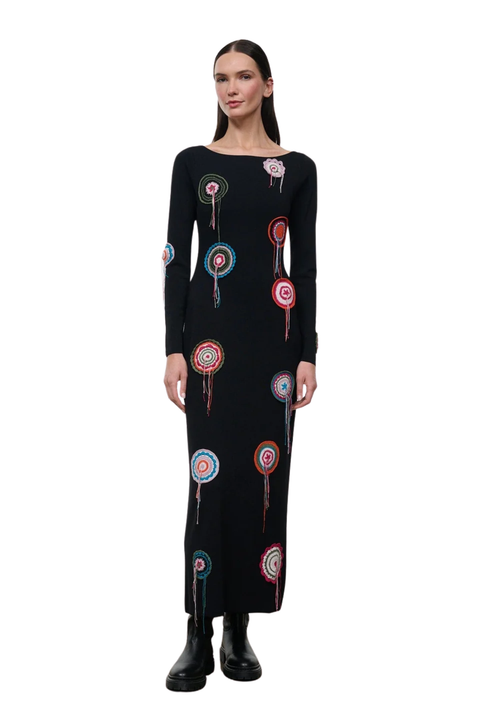 A woman modeling a long-sleeve black maxi dress with colorful circular embroidered motifs, paired with black ankle boots, against a plain white background.