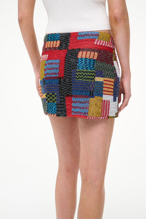 Close up back-facing woman wearing a beaded mini skirt that features a multicolor patchwork design, paired with a white tank. 