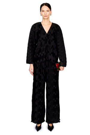 A model stands against a white studio background wearing a black textured, patterned two-piece outfit consisting of a long-sleeve V-neck top and matching wide-leg trousers, paired with black pointed shoes.