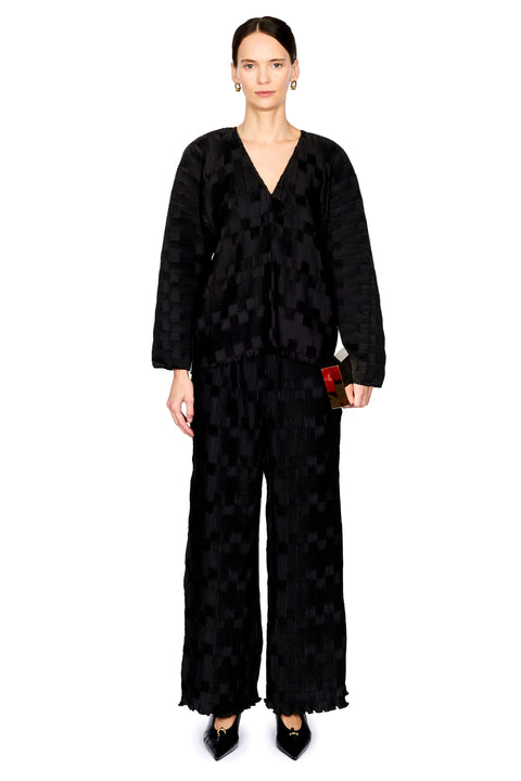 A model stands against a white studio background wearing a black textured, patterned two-piece outfit consisting of a long-sleeve V-neck top and matching wide-leg trousers, paired with black pointed shoes.
