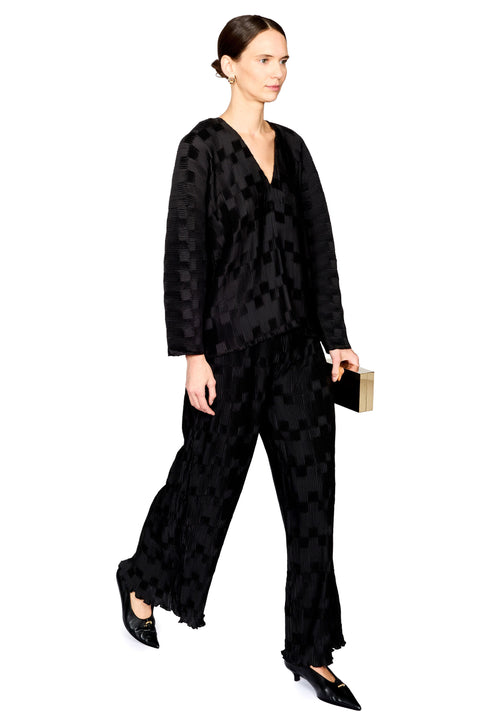 A model wearing a black patterned two-piece set consisting of a long-sleeve V-neck top and matching wide-leg trousers, holding a small black clutch with a gold edge, set against a plain white background.