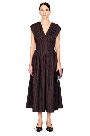 A model stands against a plain white background wearing a sleeveless dark brown midi dress with thin vertical black pinstripes, a deep V-neckline, and a gathered waist. She is holding a small black clutch and wearing black pointed flats.