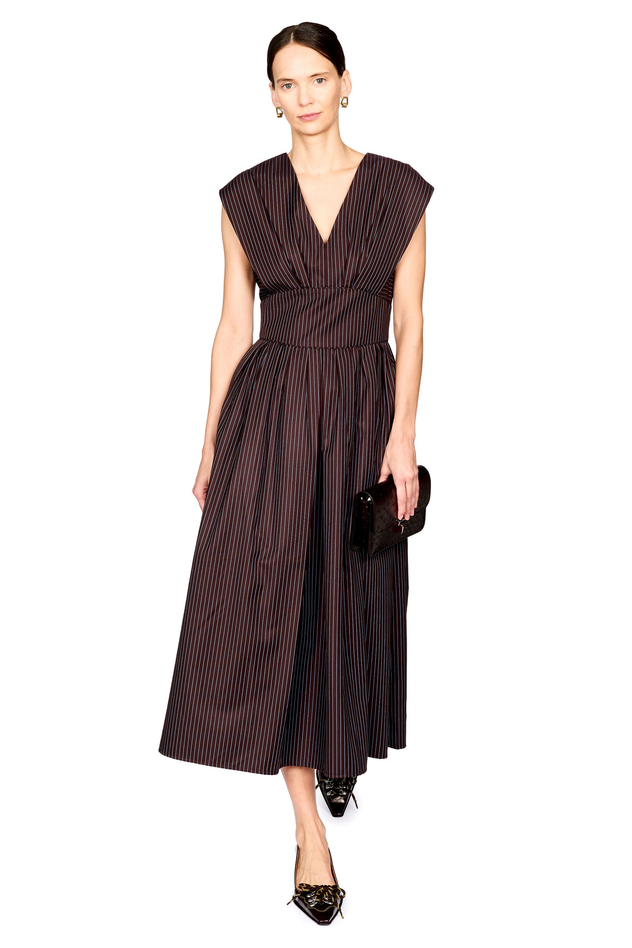 A woman modeling a sleeveless dark brown midi dress with thin vertical pinstripes, a V-neckline, and a gathered waist, paired with black pointed shoes and a small black clutch, against a plain white background.
