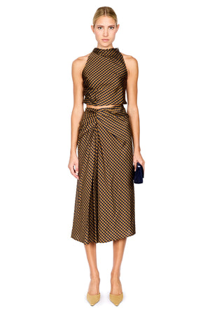 A model stands against a white background wearing a brown and navy checkered halter crop top and matching draped midi skirt, paired with beige pointed-toe heels and holding a black clutch.