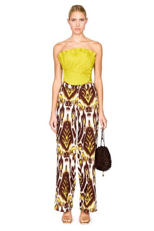 A model stands against a white background wearing a strapless bright lime top and wide-leg trousers with a brown, yellow, and white ikat pattern. She is holding a small dark brown textured handbag.