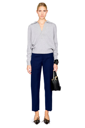 A model stands against a white background wearing a light gray wrap-style knit sweater, navy blue cropped trousers, black pointed flats, and holding a black handbag.