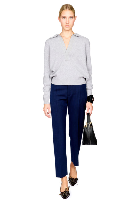A model wearing a light gray wrap-style knit sweater, navy blue cropped trousers, and black pointed flats, holding a black handbag against a plain white background.