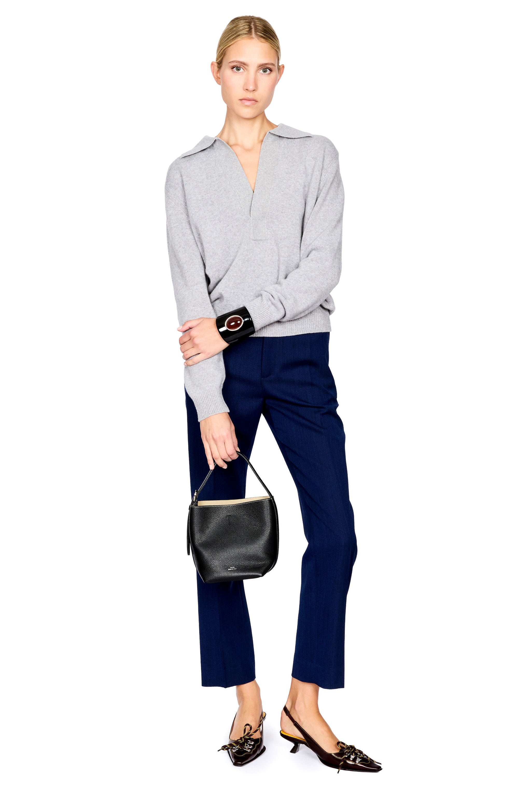 A model stands against a white background wearing a light gray half-zip sweater, navy blue cropped trousers, and black pointed flats, while holding a black leather handbag.