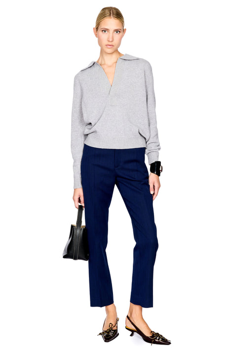 A model stands against a white background wearing a light gray wrap-style knit top, navy blue cropped trousers, and black pointed flats, while holding a small black handbag.