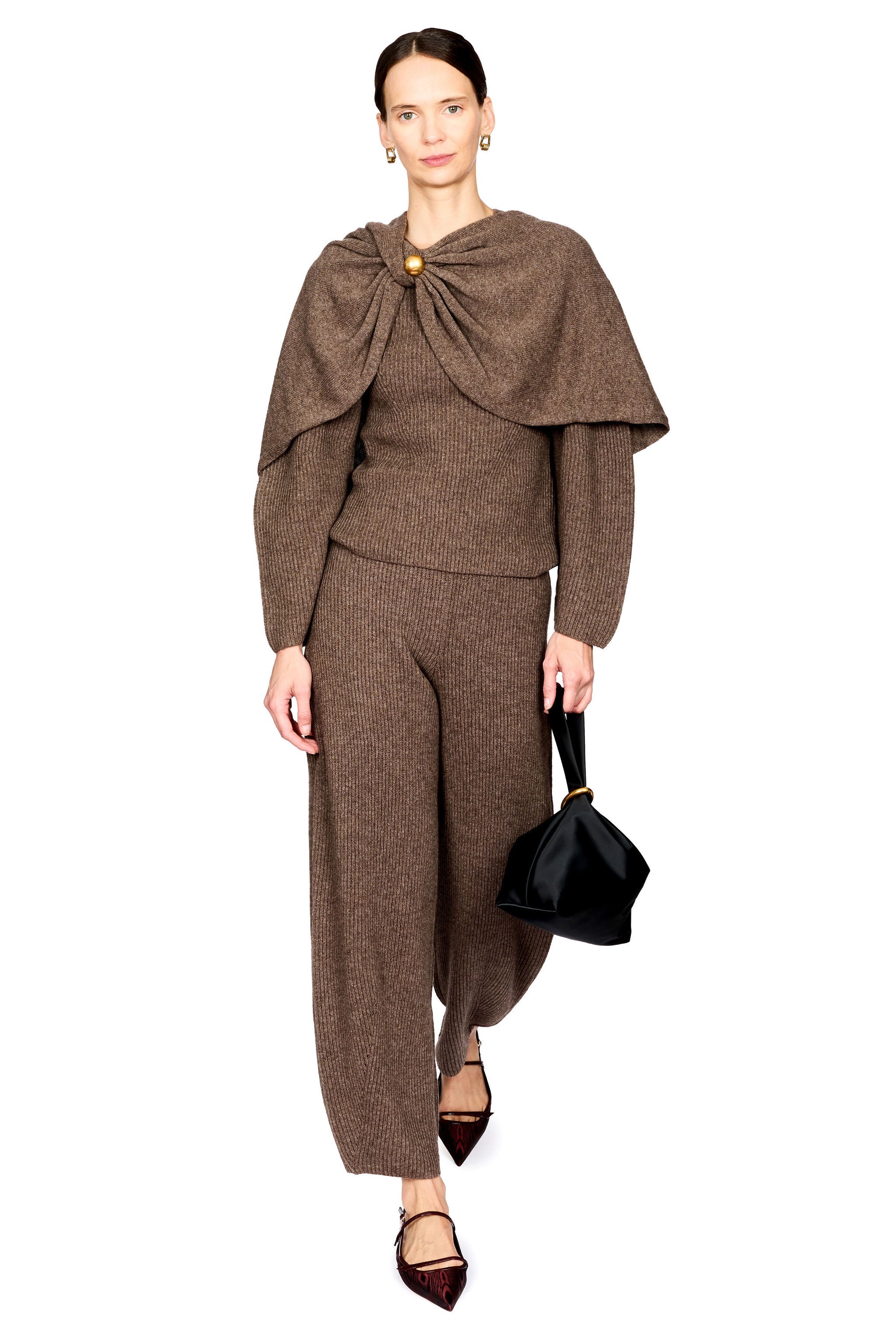 A model wearing a brown knit matching set consisting of a long-sleeve top with a draped collar and broch, and matching trousers, styled with dark pointed flats and a black handbag against a white background.