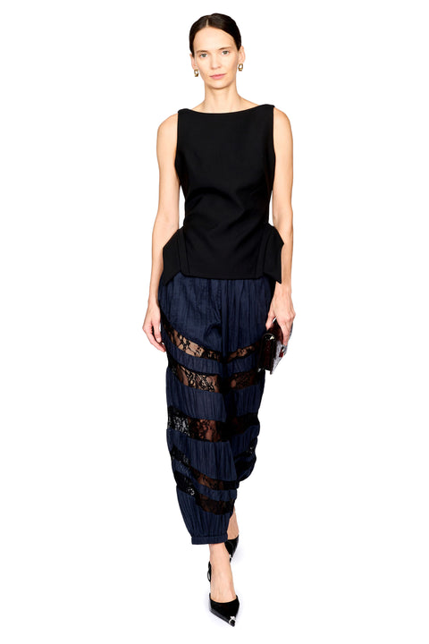 A model wearing a sleeveless black peplum top and dark navy pleated trousers with sheer black lace bands, standing against a plain white background.