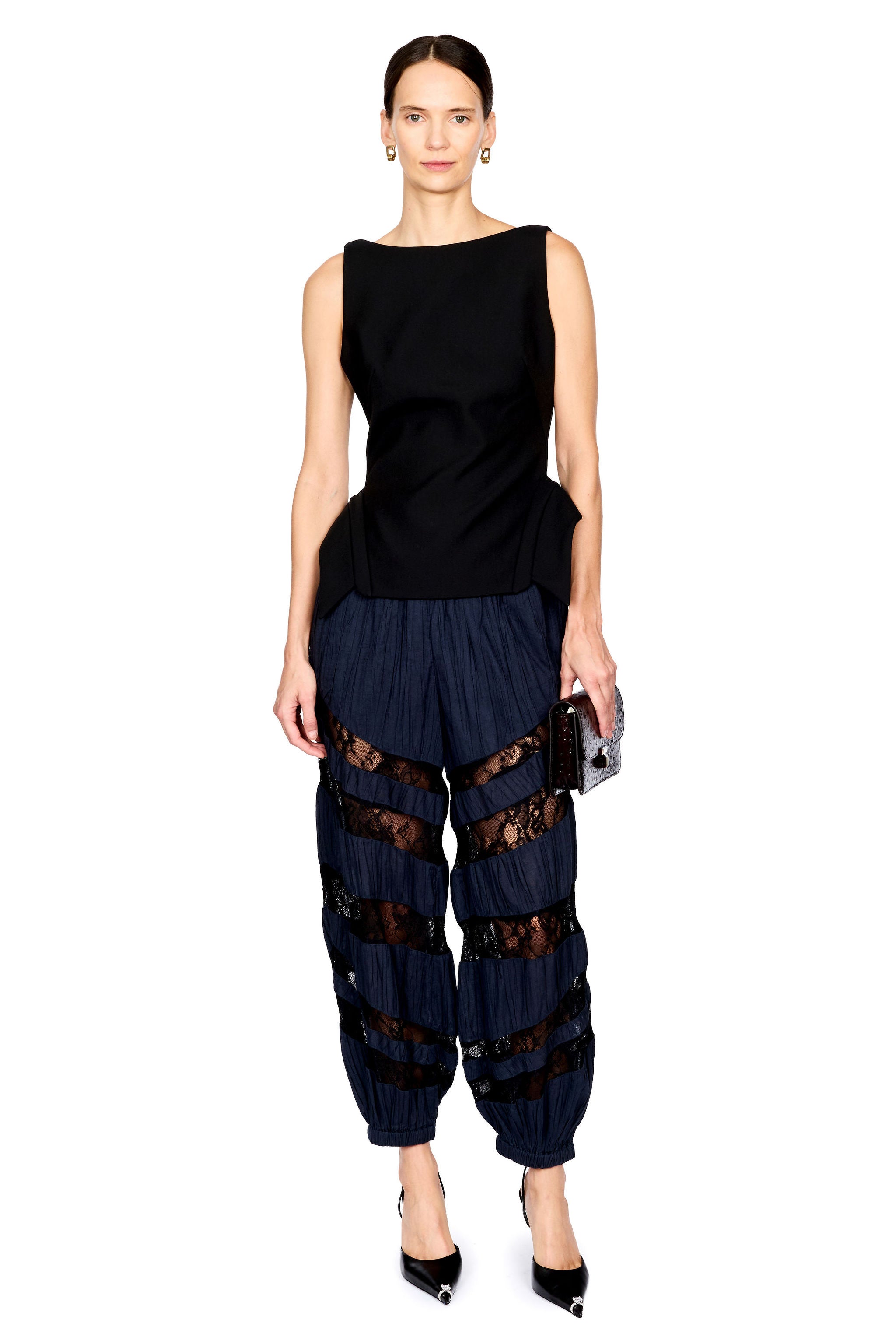 A model wearing a sleeveless black top and navy blue pleated harem-style pants with sheer black lace panels, holding a small brown clutch against a plain white background.
