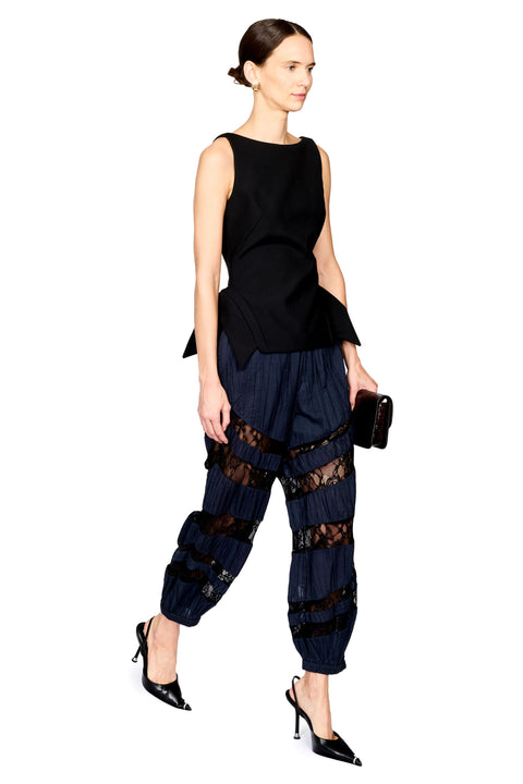 A model is shown in profile wearing a sleeveless black peplum top and dark navy pleated harem-style pants with sheer black lace panels, holding a small brown clutch against a plain white background.