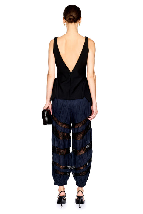 A model shown from the back wearing a black sleeveless top with a deep V-back and dark pleated harem-style pants with sheer black lace panels, against a plain white background.