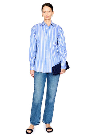 A woman modeling a light blue and white vertically striped button-down shirt with blue jeans, black slide sandals, and a dark navy clutch, set against a plain white background.