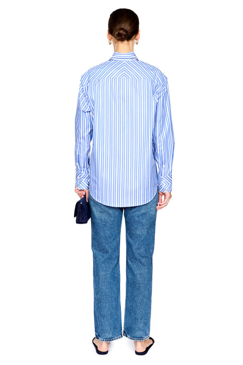 A woman viewed from the back wearing a light blue and white vertically striped button-down shirt, blue jeans, and black sandals, holding a small dark blue clutch against a plain white background.