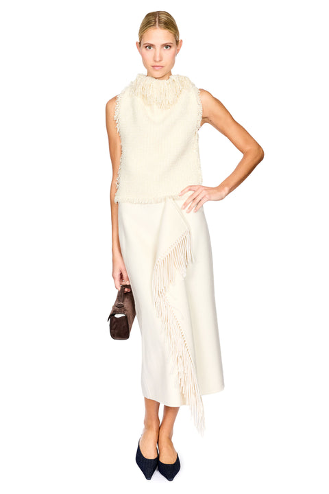 A model wearing a sleeveless cream knit dress with a high ruffled collar and fringe detailing, paired with dark navy pointed-toe heels and holding a small brown handbag against a solid white background.