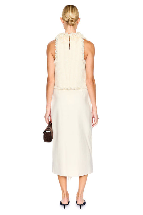 A woman shown from the back wearing a sleeveless cream top with a ruffled high neckline and a matching skirt. She is holding a small dark brown handbag and wearing black pointed-toe heels against a plain white background.