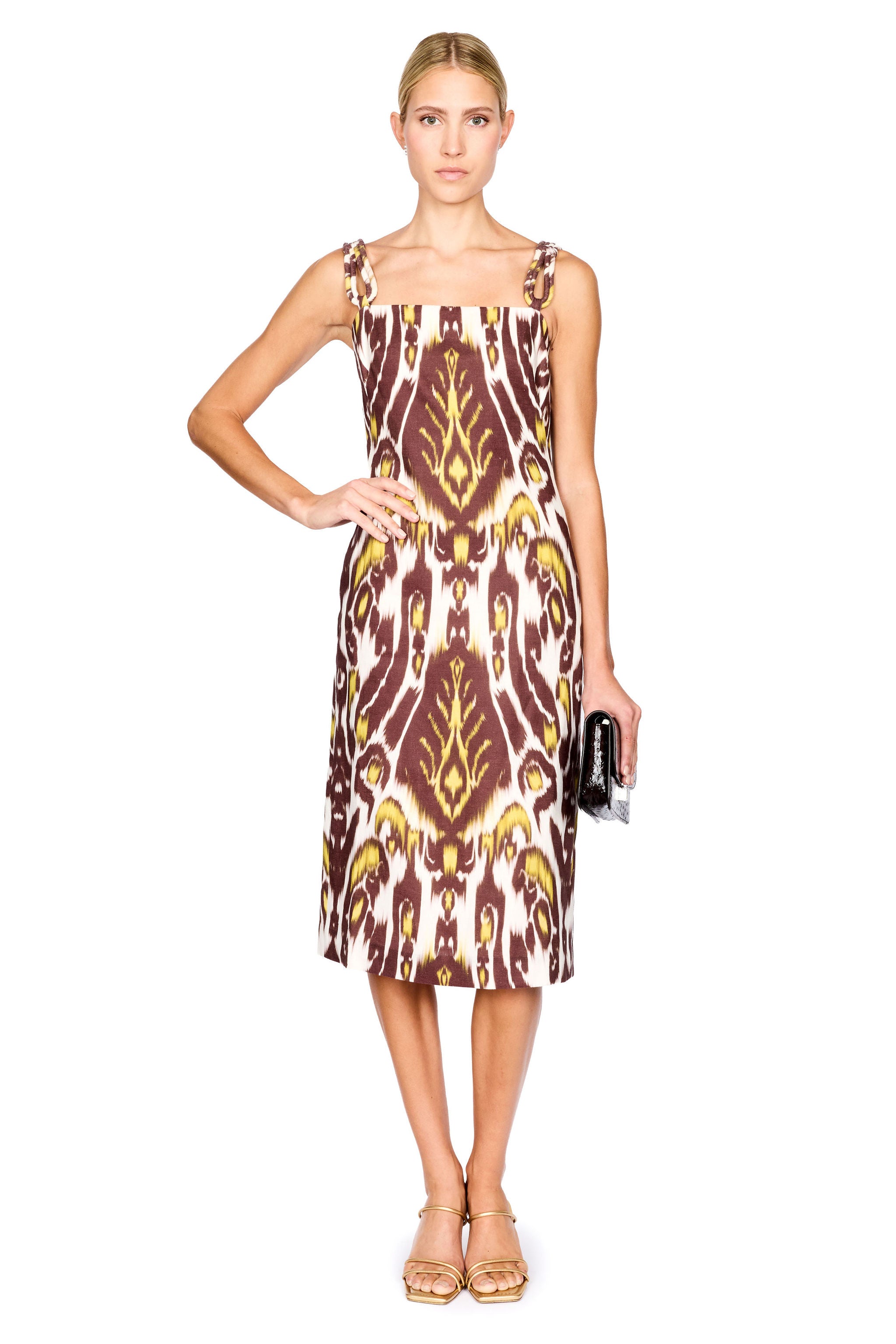 A woman modeling a midi dress with a brown, gold, and cream ikat pattern, paired with gold sandals and holding a small black clutch, set against a plain white background.