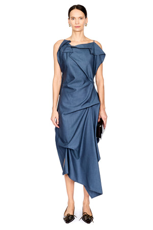 A model wearing a slate blue one-shoulder draped midi dress with a high-low asymmetrical hem, paired with black pointed flats and holding a small black clutch, set against a plain white background.