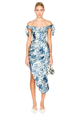 A model wearing an off-the-shoulder midi dress with a blue and white tropical floral print, holding a matching patterned clutch and wearing white pointed-toe heels against a plain white background.