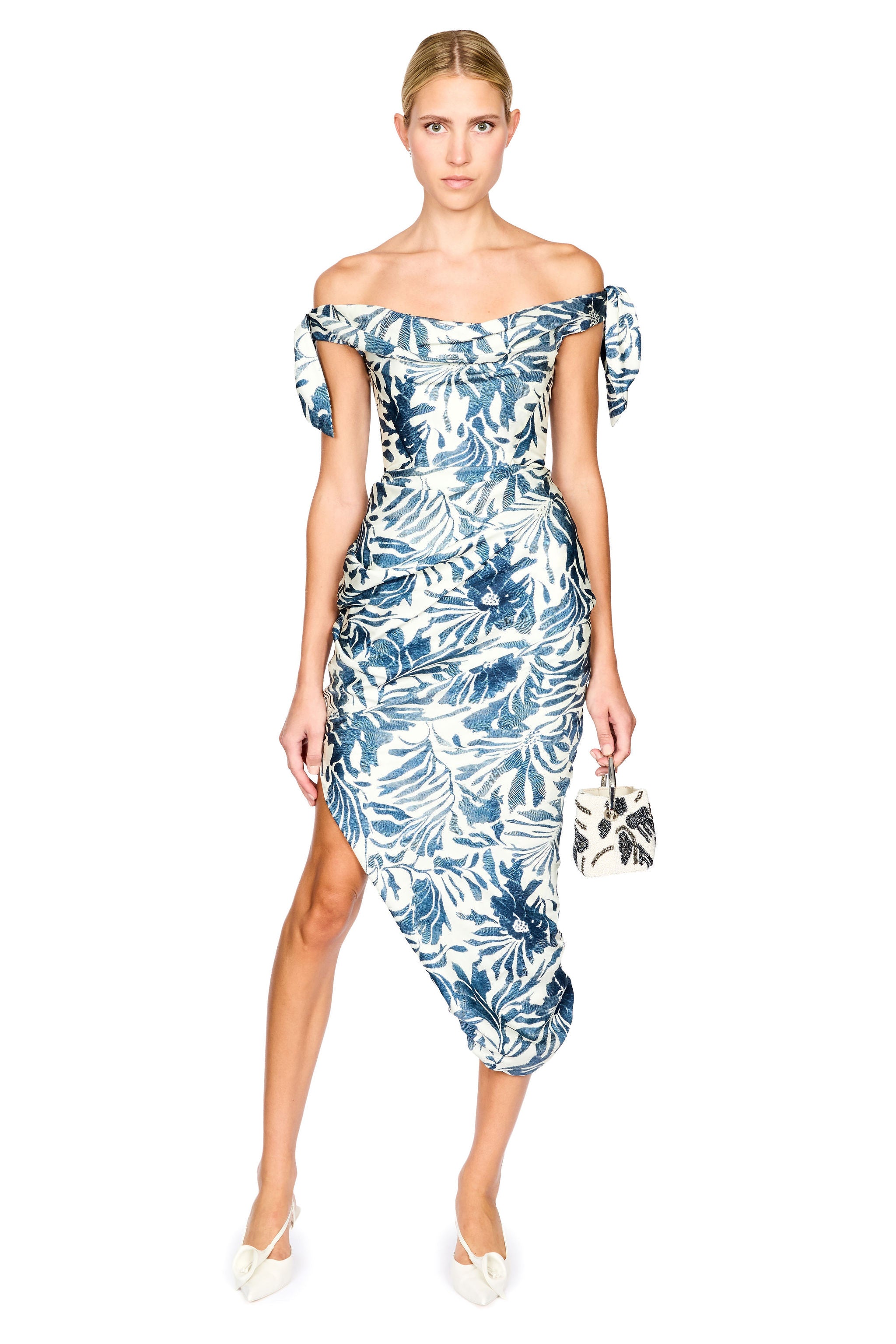 A woman modeling an off-the-shoulder midi dress with a blue and white tropical leaf pattern, paired with white pointed-toe heels and a small patterned handbag, set against a plain white background.