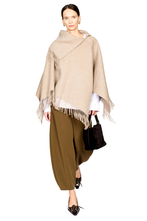 A model wearing a beige fringed wrap poncho over a white top and olive green wide-leg pants, holding a black handbag against a plain white background.
