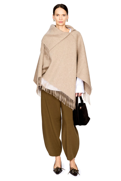 A model stands against a white studio background wearing a beige shawl poncho with fringe, olive green wide-leg pants, black pointed flats, and holding a black handbag.