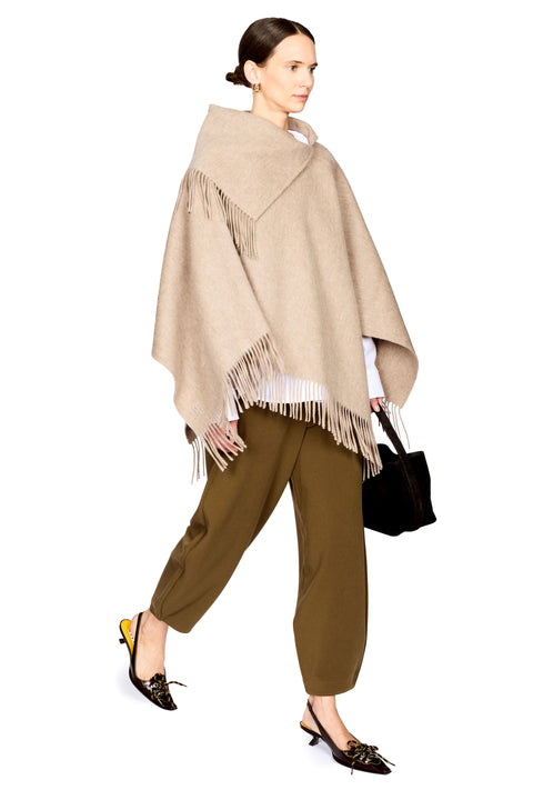 A woman modeling a beige fringed wrap poncho over a white top and olive green cropped pants, holding a black handbag against a plain white background.