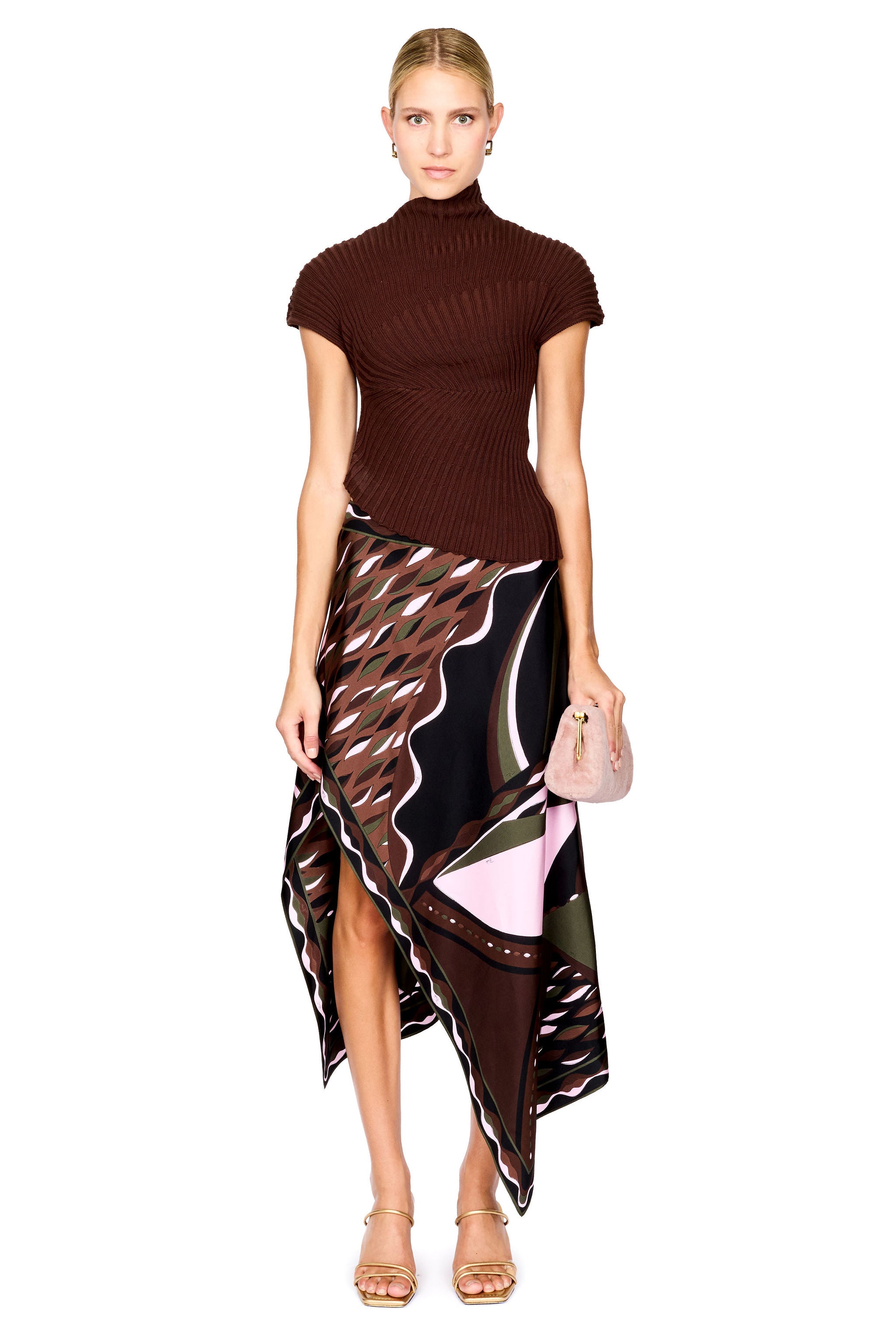 A model stands against a white studio background wearing a dark brown ribbed short-sleeve mock-neck top and a high-low wrap skirt with a black, brown, beige, and pink abstract print. She is holding a small blush pink handbag and wearing gold high-heeled sandals.