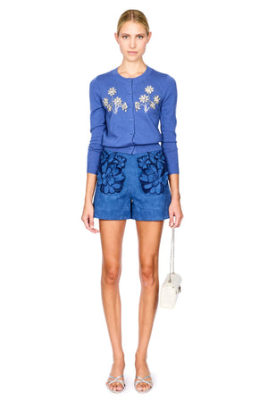 A model stands against a white studio background wearing a blue knit cardigan with gem floral embroidery and matching blue shorts with dark blue floral applique. She is holding a small white handbag and wearing silver high-heeled sandals.
