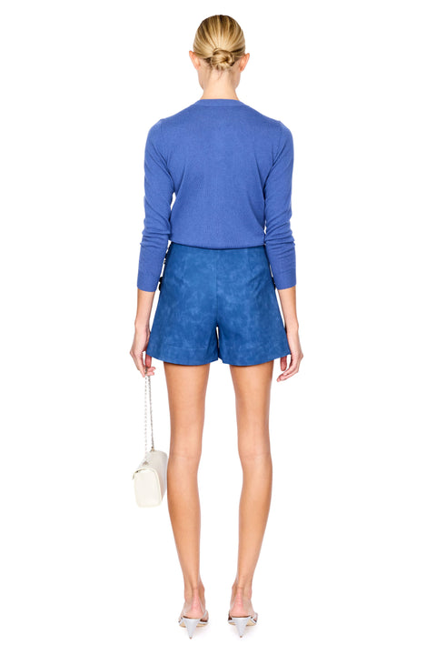 A model stands against a white studio background wearing a blue knit cardigan and matching blue shorts. She is holding a small white handbag and wearing silver high-heeled sandals.