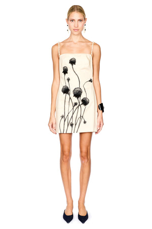 A model wearing a cream-colored spaghetti strap mini dress with a black dandelion print, paired with black pointed-toe heels against a solid white background.
