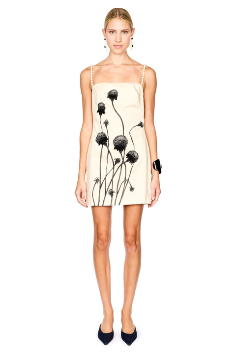 A model wearing a cream-colored spaghetti strap mini dress with a black dandelion print, paired with black pointed-toe heels against a solid white background.