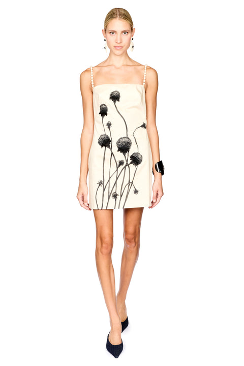 A model wearing a cream-colored spaghetti strap mini dress with a black dandelion print, styled with black pointed-toe heels and a black wrist accessory against a solid white background.