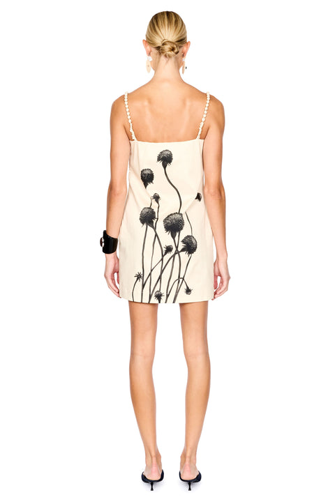 A woman viewed from the back wearing a cream-colored spaghetti-strap dress with a black dandelion print, paired with black high heels and a black bracelet, against a solid white background.