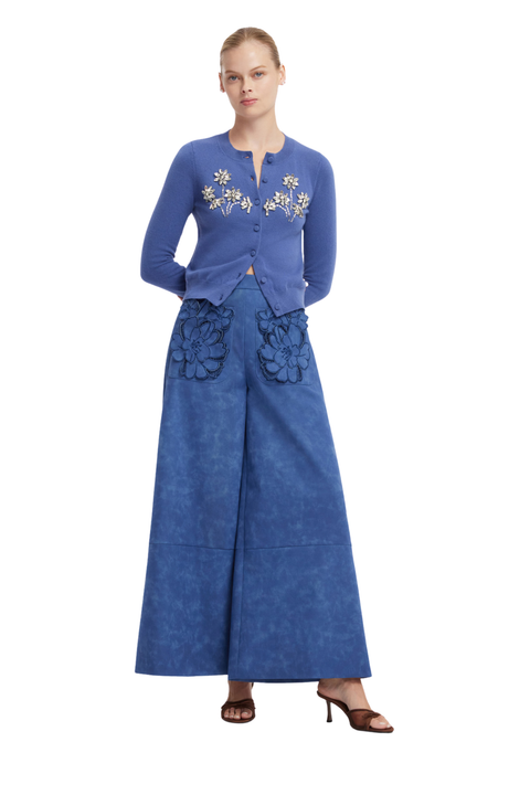 A model wearing a blue two-piece outfit consisting of a long-sleeve button-front cardigan with white floral embroidery and matching wide-leg trousers with dark floral applique, paired with brown heeled sandals against a white background.