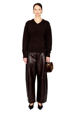 A model stands against a white background wearing a dark brown v-neck knit sweater, wide-leg dark brown faux-leather pants, black pointed shoes, and holding a small brown fur handbag.